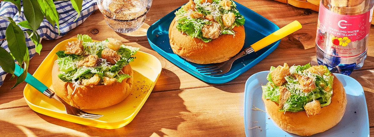 Three caesar salads in bread bowls on colourful plates on a picnic table