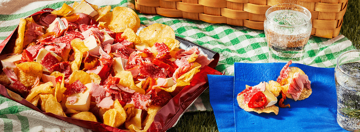 Potato chip charcuterie served in a kettle chip bag topped with brie, and deli meats. Two chips on a napkin to the side.