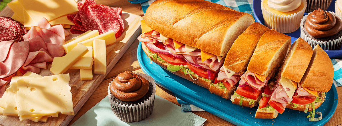 Classic picnic set up with Dagwood sandwich, and charcuterie board to the side. Chocolate cupcake on a plate in the foreground.